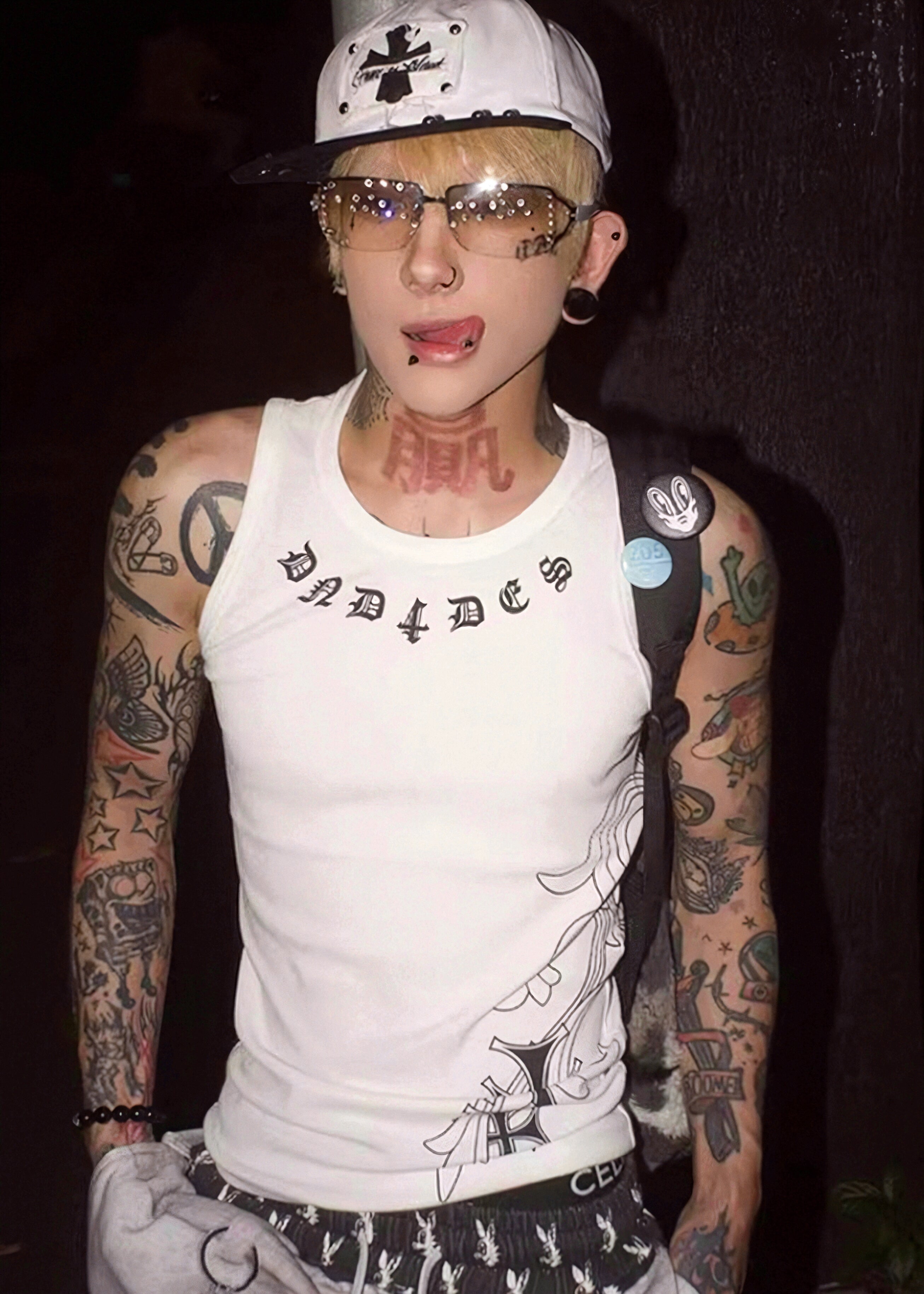 Person wearing a white tank top with tattoos and a cap, standing against a dark background.